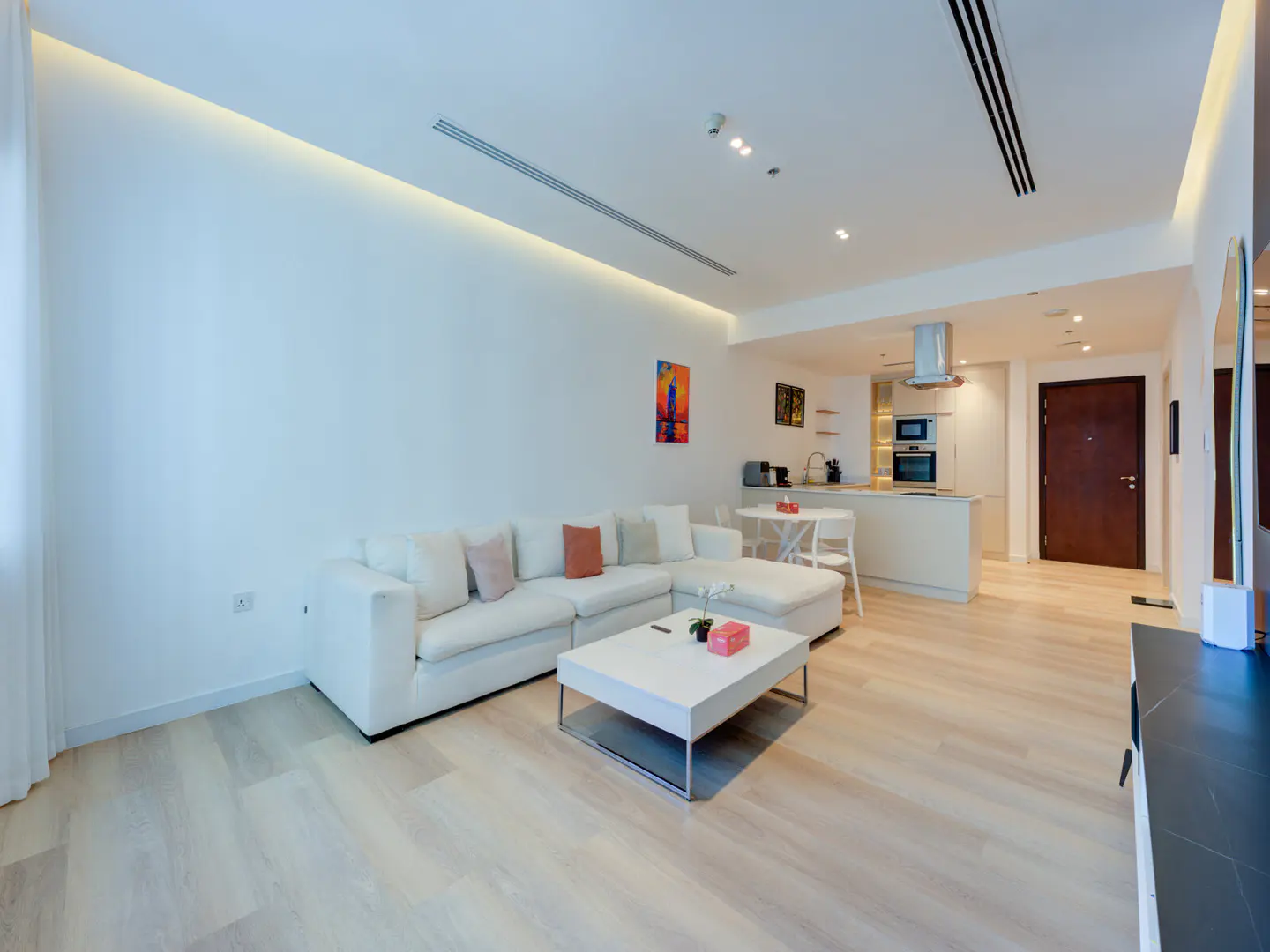 Bright, modern apartment interior with white sectional sofa, coffee table, and open kitchen. Light wood floors and minimalist decor.