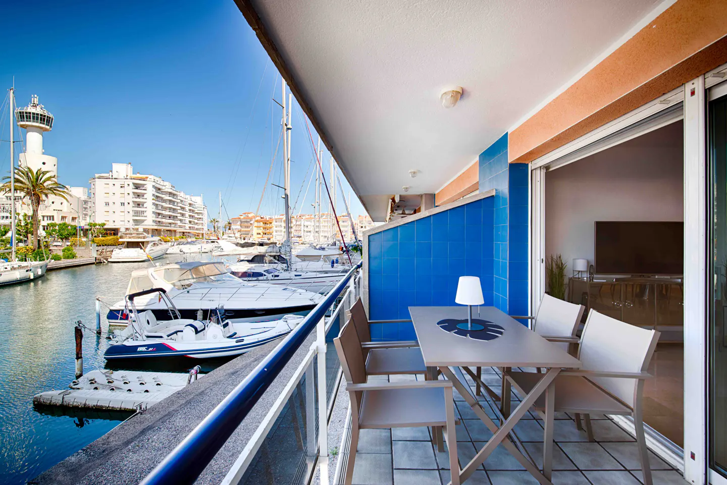 Balcony view of a marina with boats. Table and chairs on the balcony. Blue tiled wall and sliding glass doors.