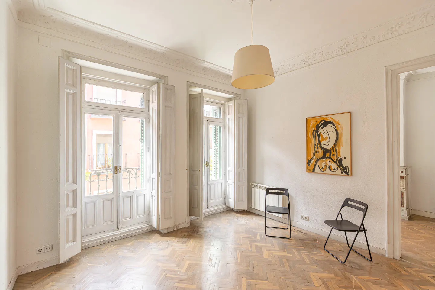 Bright, empty room with herringbone wood floors, white walls, and two black folding chairs. Two sets of white doors lead to balconies. A painting hangs on the wall.