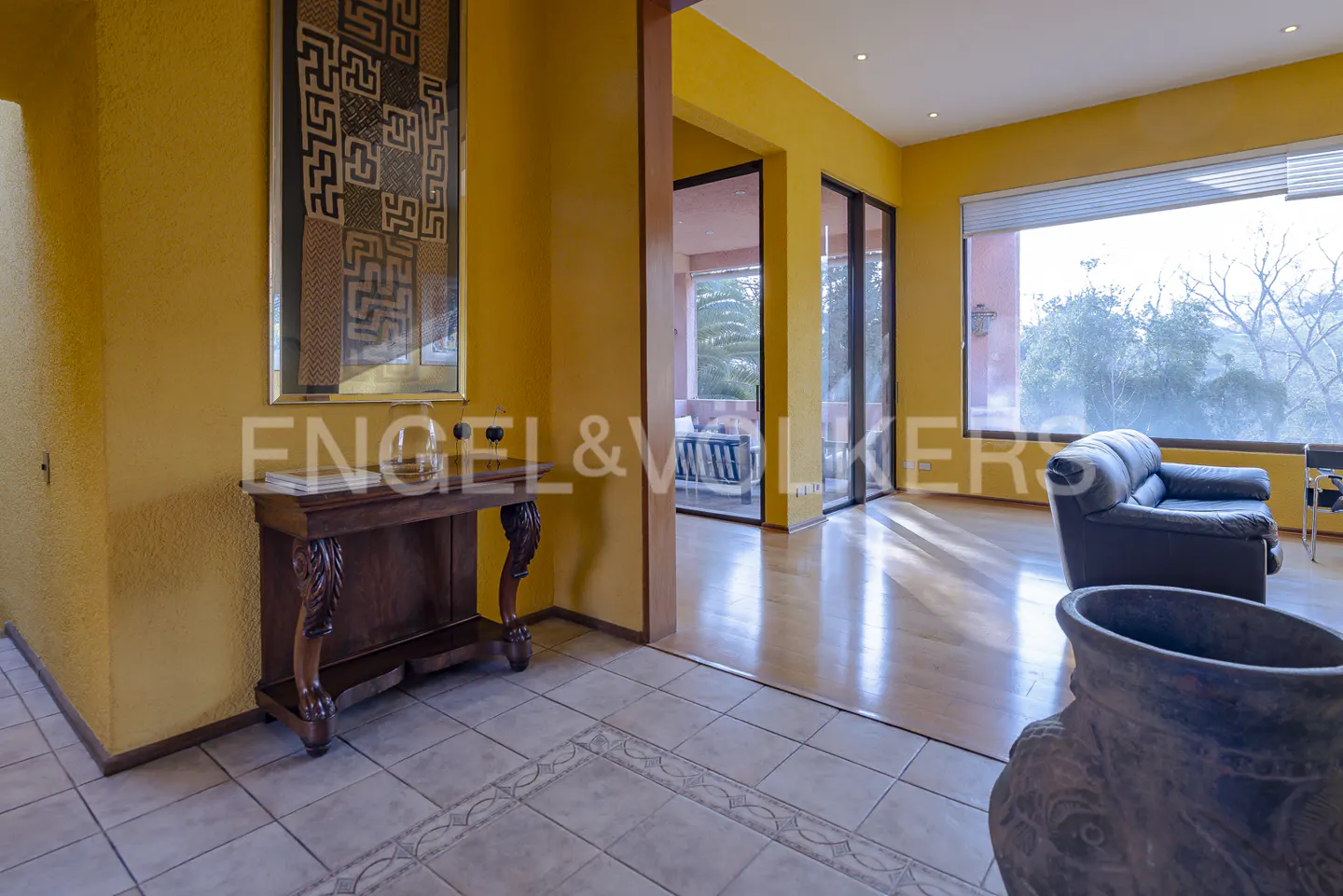 Interior view of a yellow-walled home with a dark wood table, art, and a glimpse of a living room with a view.