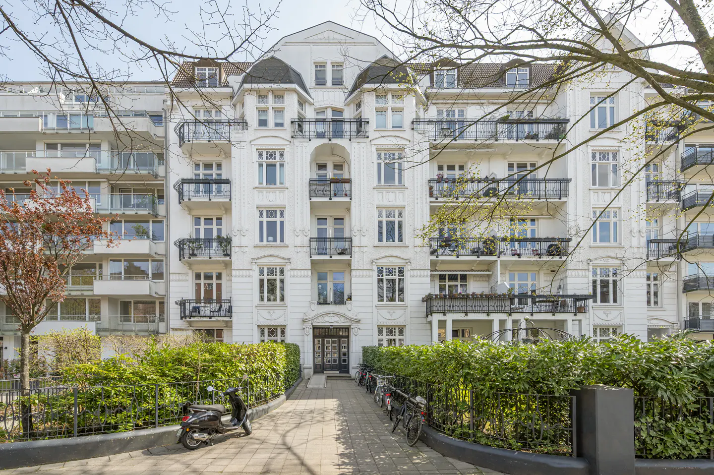 Exterior view of a white apartment building with balconies, a hedge, and parked bikes.