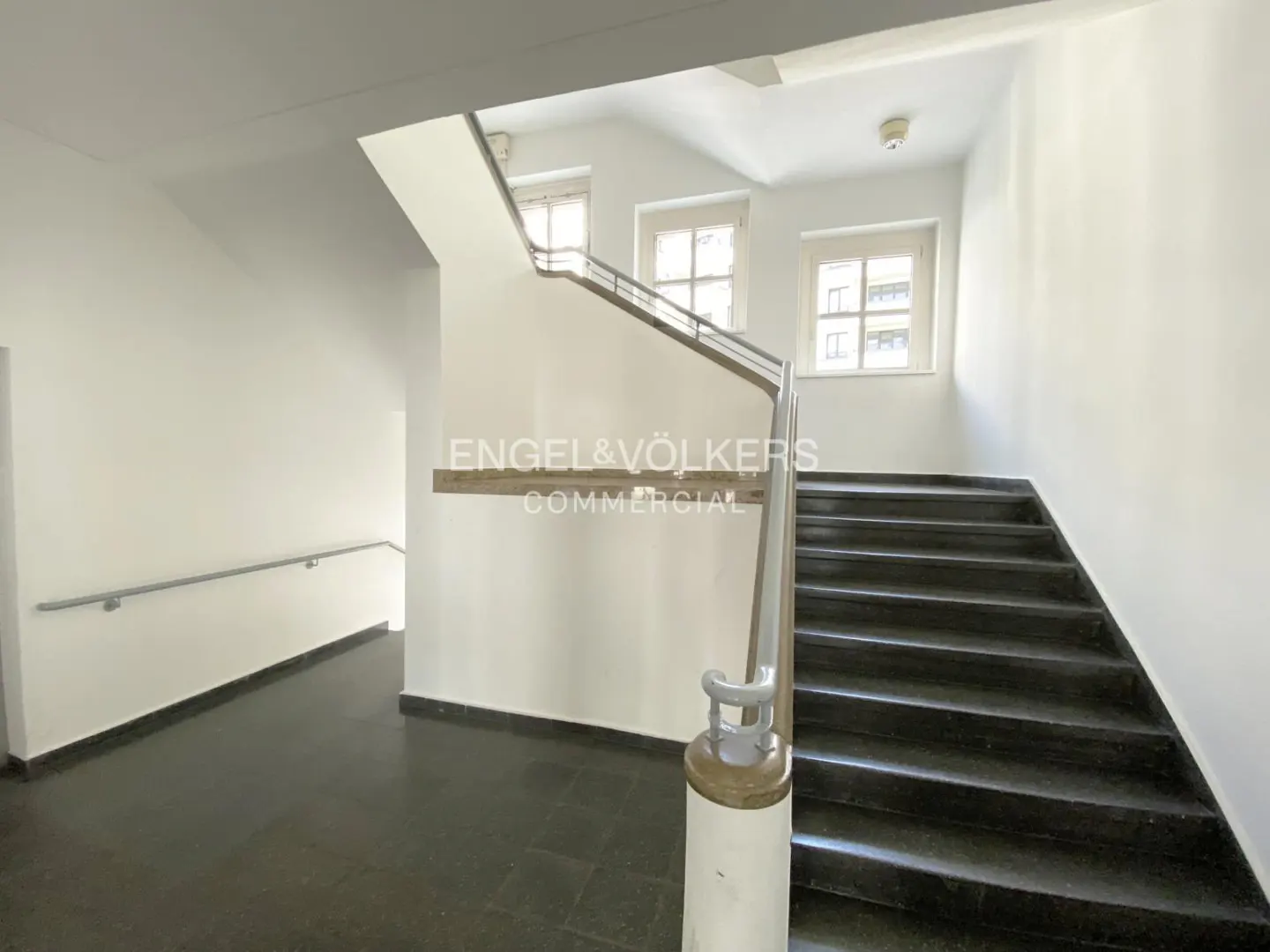 Interior view of a stairwell with black stairs, white walls, and a metal handrail. Windows are visible on the upper level. "Engel & Völkers Commercial" logo on the wall.