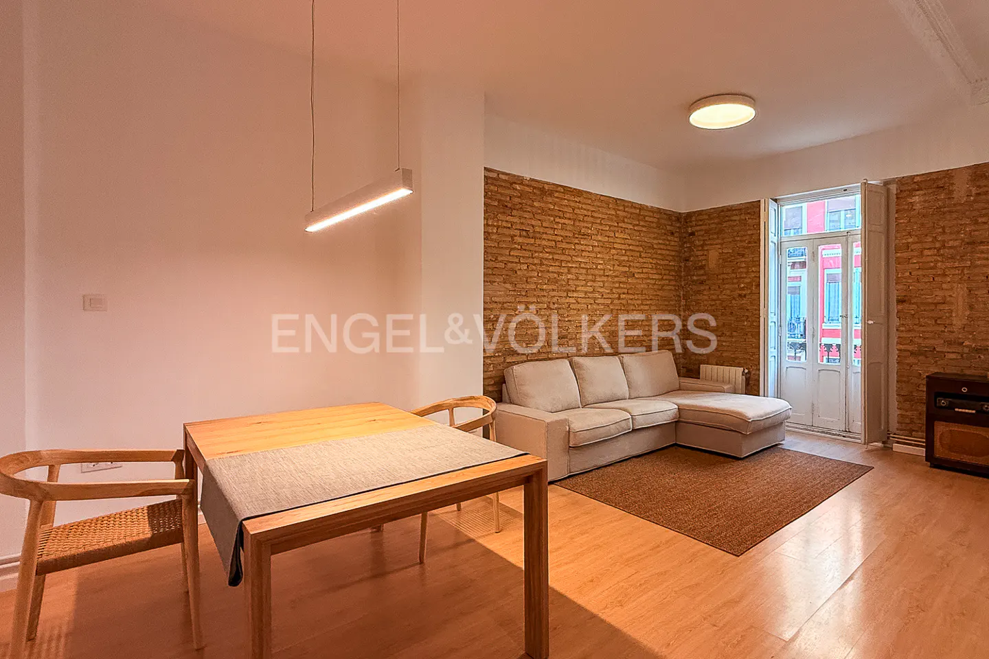A bright living room with a wooden table and chairs, a beige sofa, and a brick wall.