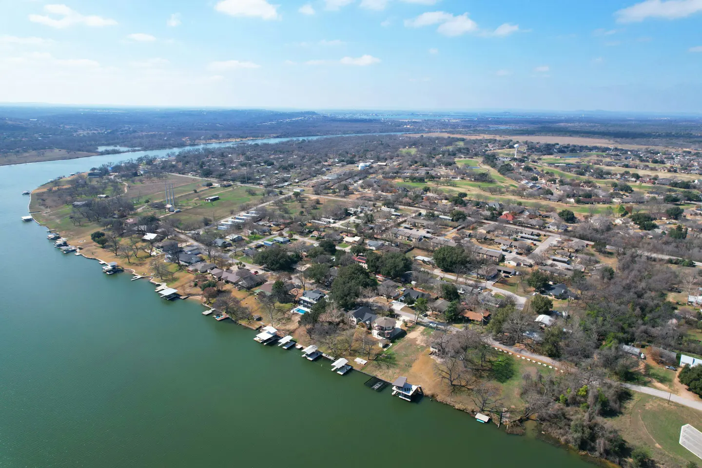 Aerial view of a lakeside neighborhood with houses, docks, and green water under a blue sky.