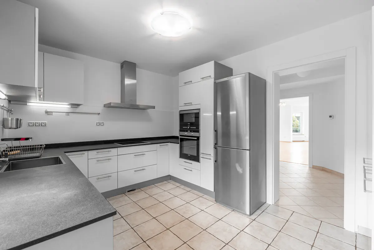 Bright kitchen with white cabinets, stainless steel appliances, and tan tile floor. Doorway leads to a room with wood floors.