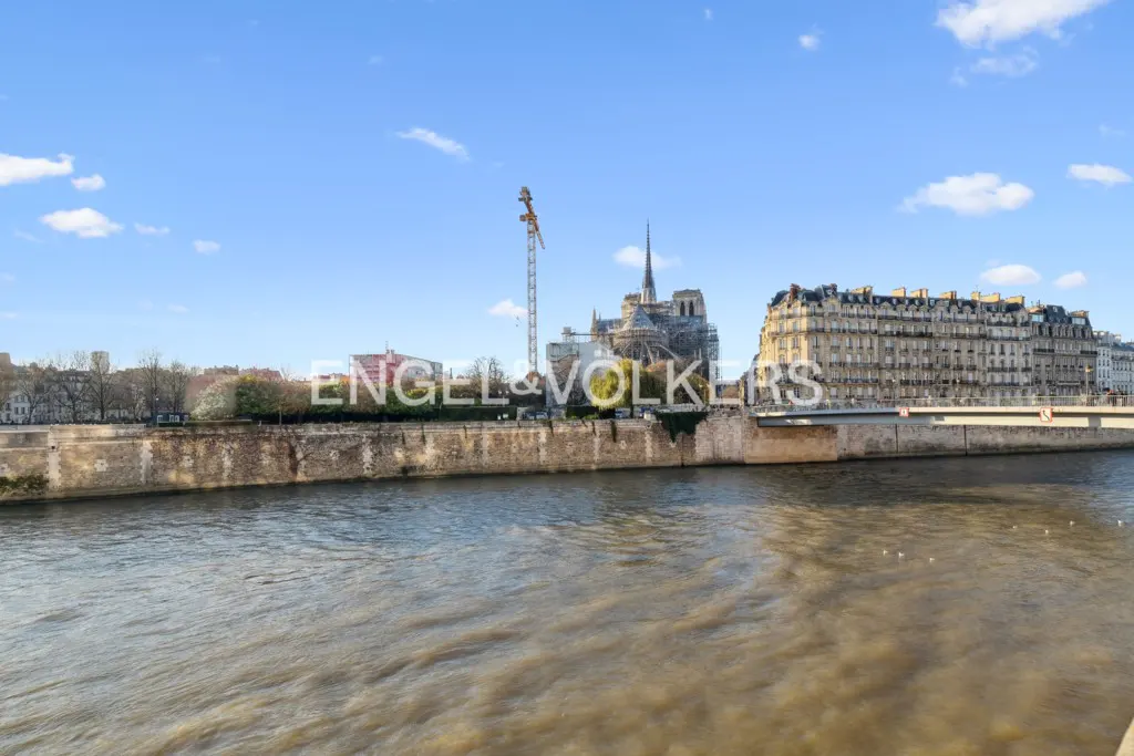 View of the Seine River in Paris with Notre-Dame Cathedral, a construction crane, and historic buildings under a blue sky.