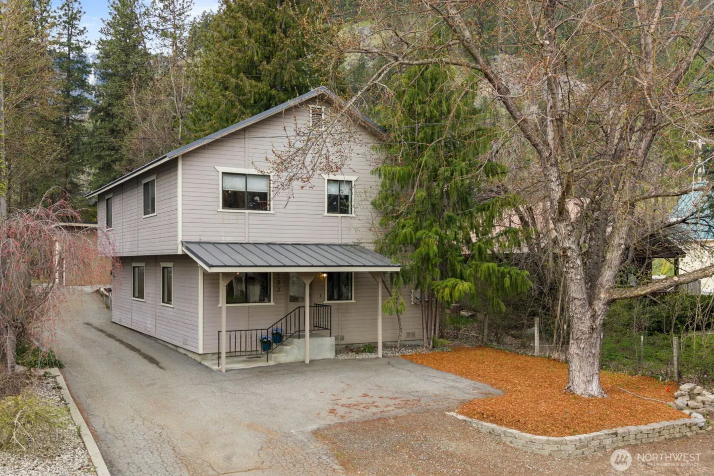 Two-story gray house with a metal roof and covered porch, surrounded by trees and a driveway.