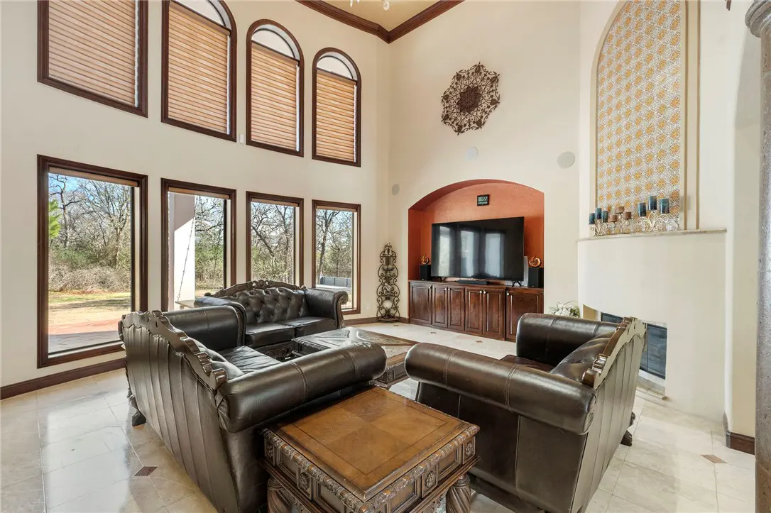 A living room with high ceilings, large windows, and brown leather furniture. A fireplace and TV are visible.