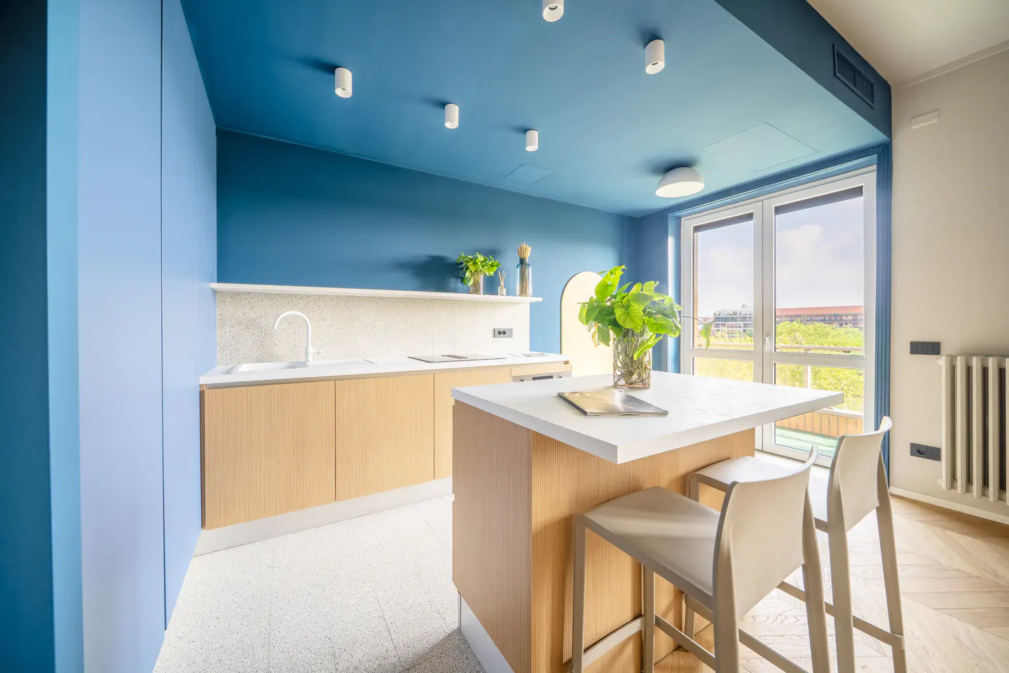 Bright kitchen with blue walls and ceiling, light wood cabinets, white countertops, island with stools, and a window view.