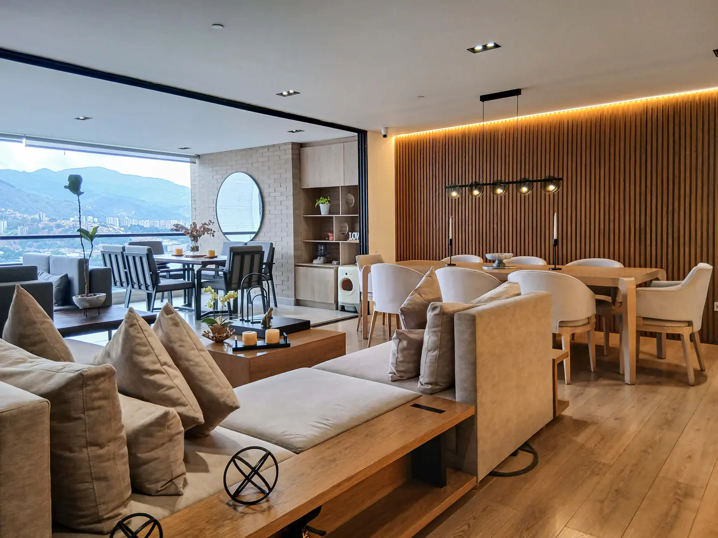 A modern living room with a beige sofa, wooden floors, and a dining area with a mountain view.