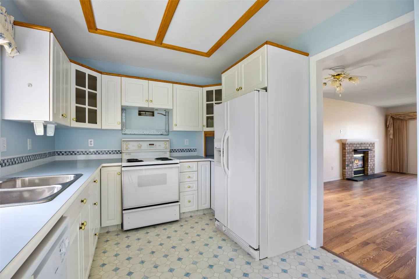 A kitchen with white cabinets, appliances, and blue accents, leading to a living room with a fireplace.