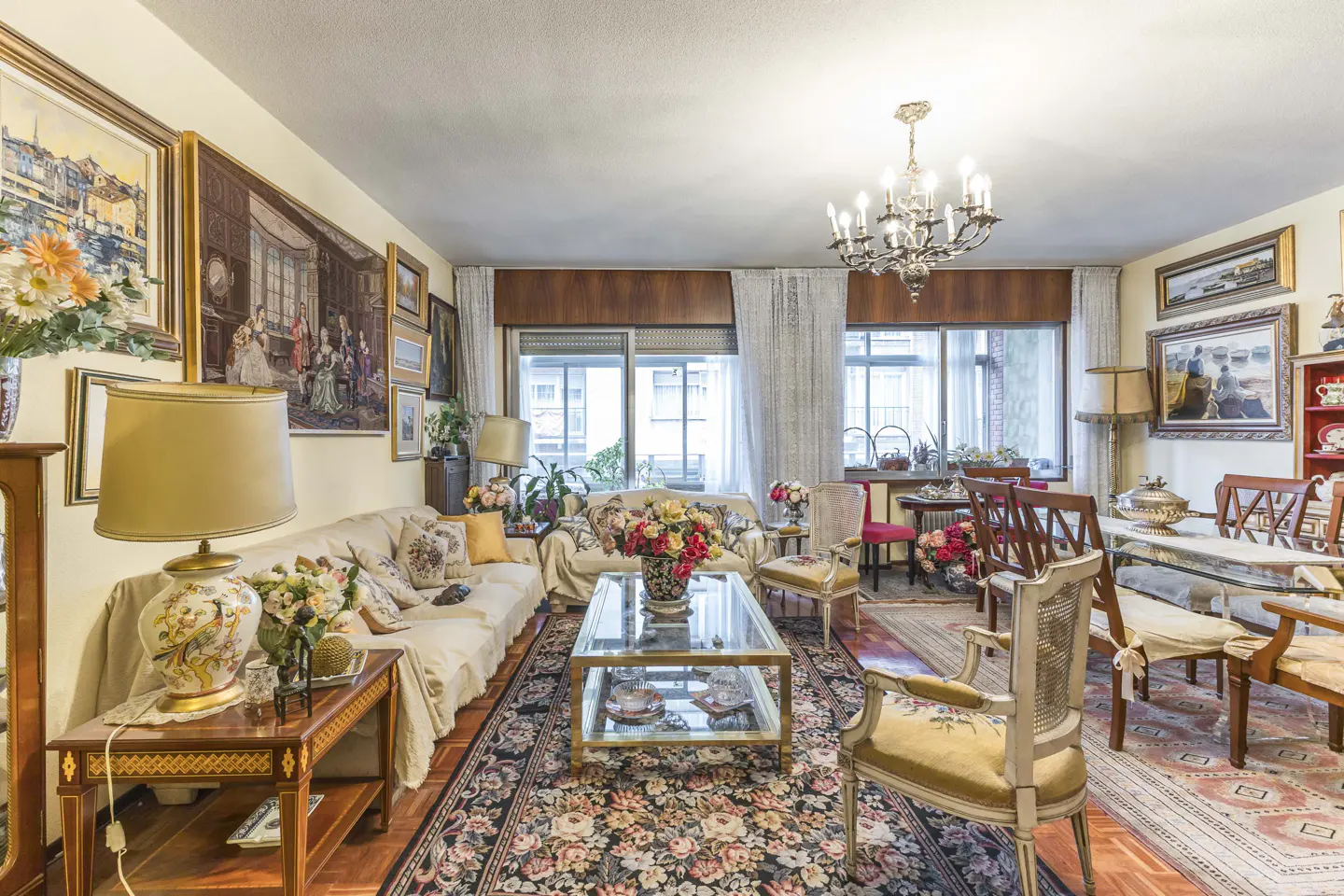 A living room with sofas, chairs, a glass coffee table, and a floral rug. Paintings adorn the walls, and a chandelier hangs from the ceiling.