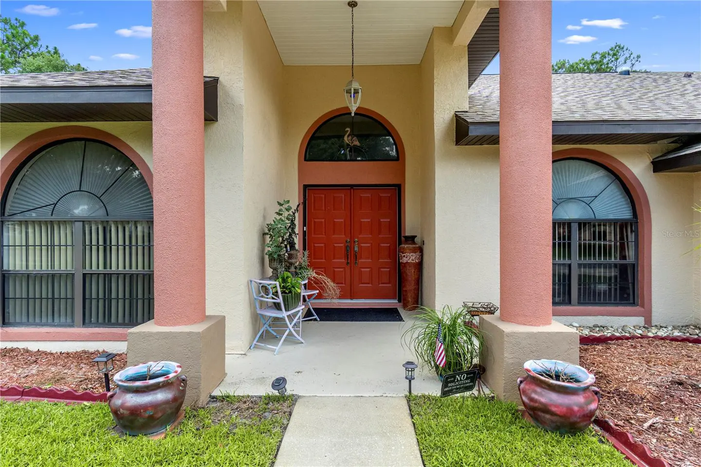 Front exterior of a beige house with red double doors, pink columns, arched windows, and potted plants.