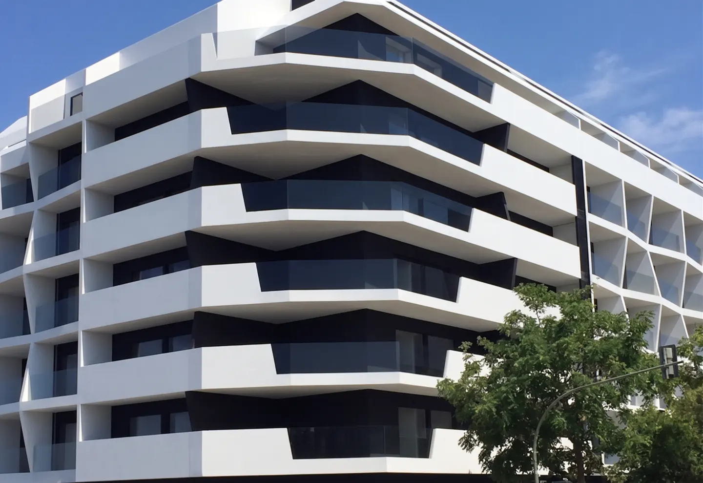 Modern white building with black accents and glass balconies under a blue sky, partially obscured by a green tree.