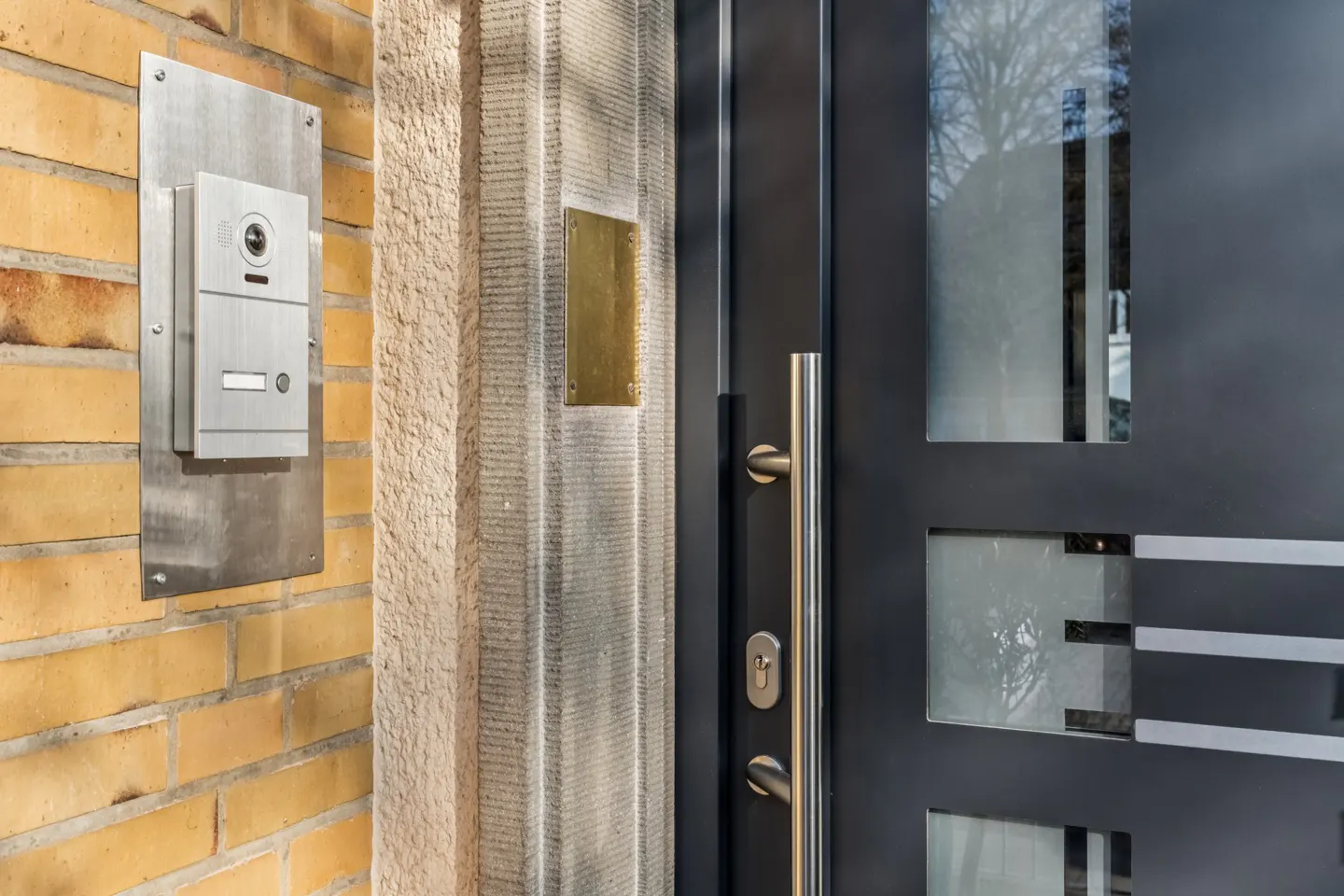 Close-up of a modern front door with a stainless steel handle, a video intercom on a brick wall, and glass panels.