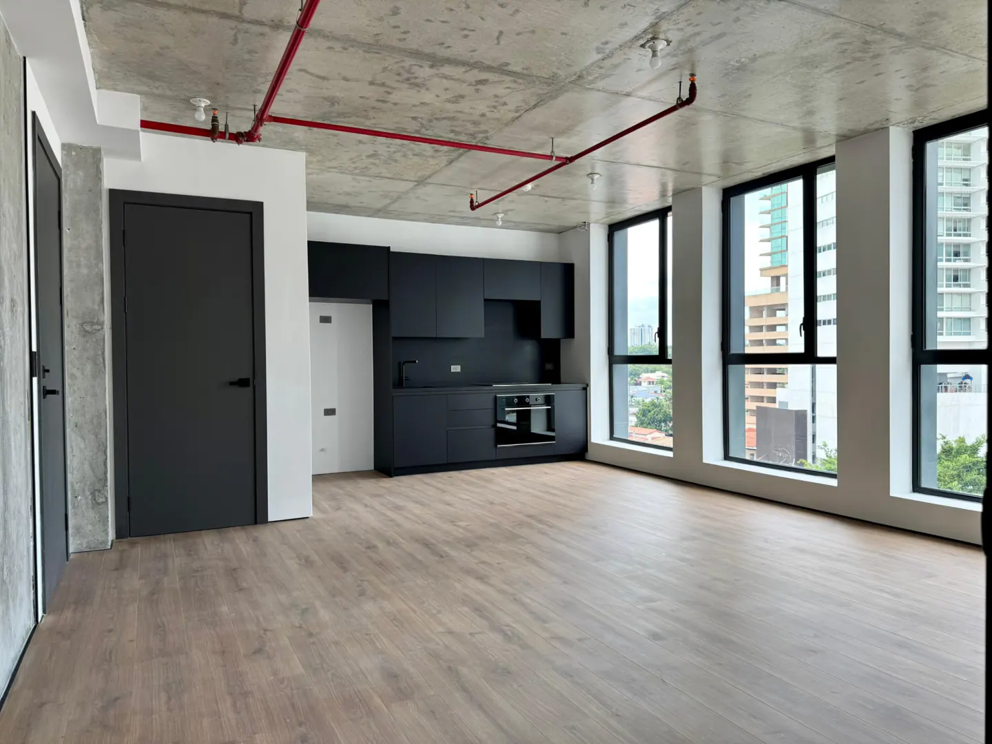An empty apartment with wood floors, black kitchen cabinets, and large windows with a city view. Red pipes run across the concrete ceiling.