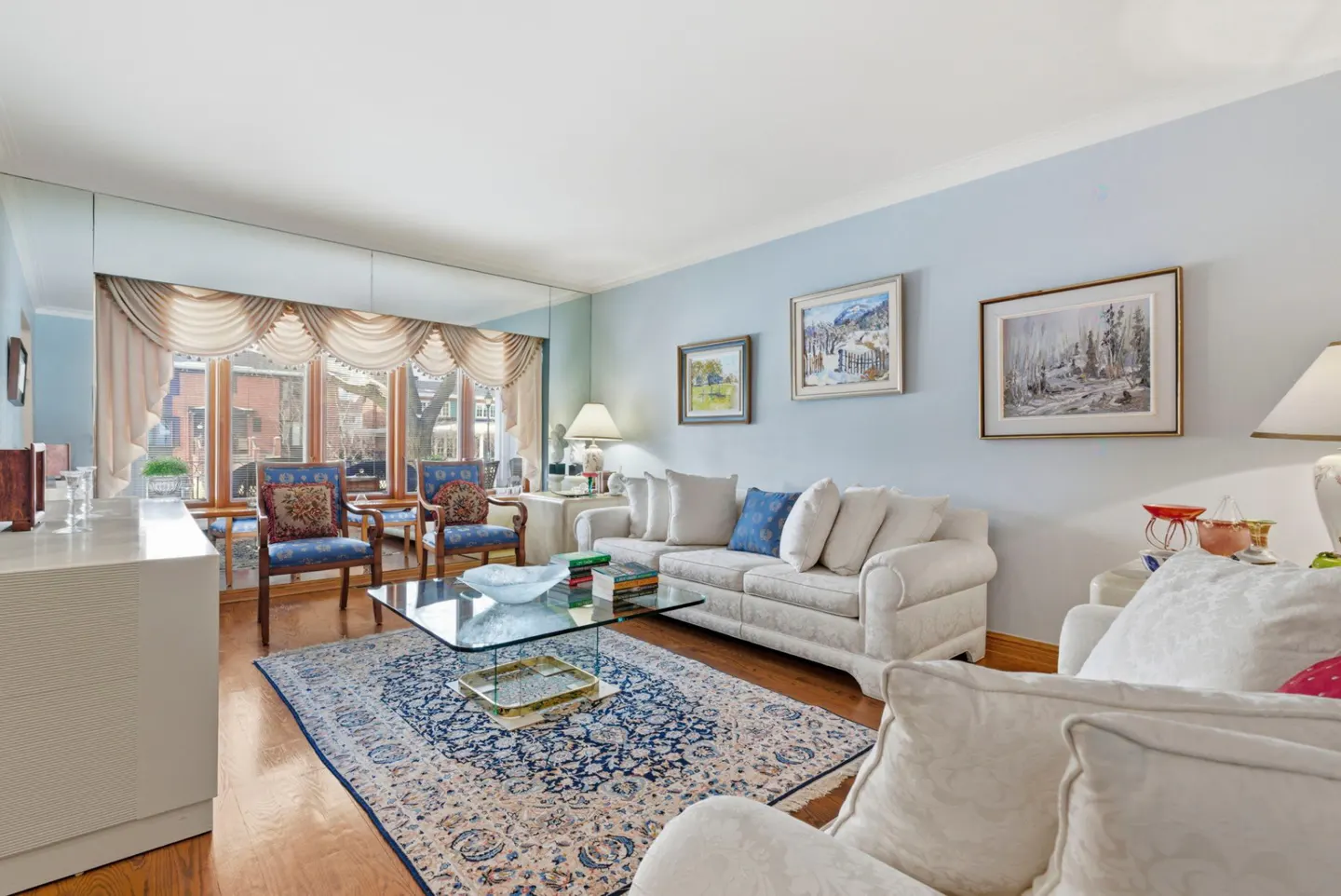 Living room with light blue walls, hardwood floors, and a large window. Two white sofas face each other over a blue patterned rug and glass coffee table.