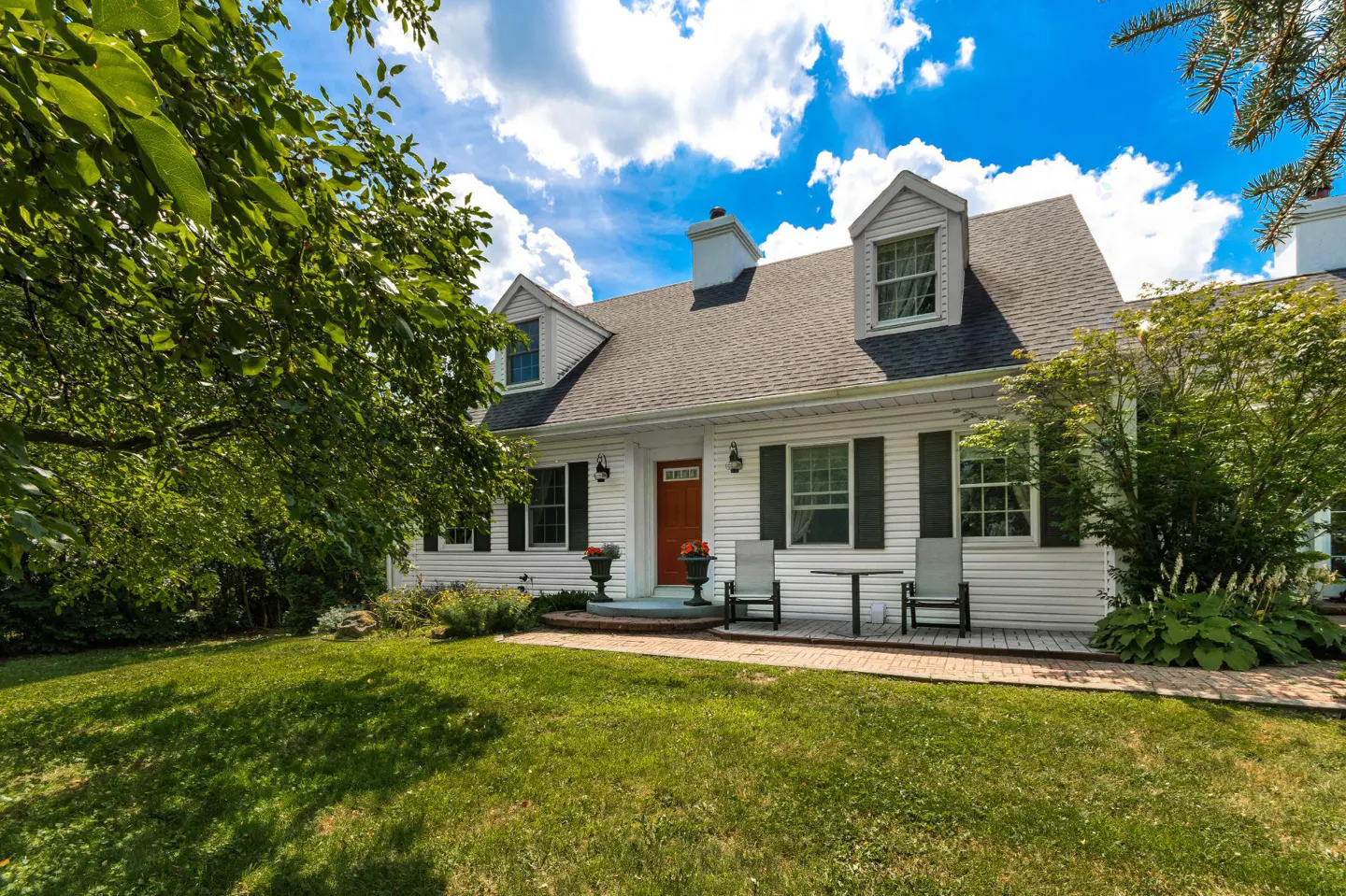 White house with black shutters and an orange door, surrounded by green trees and grass. A table and chairs sit on the front porch.
