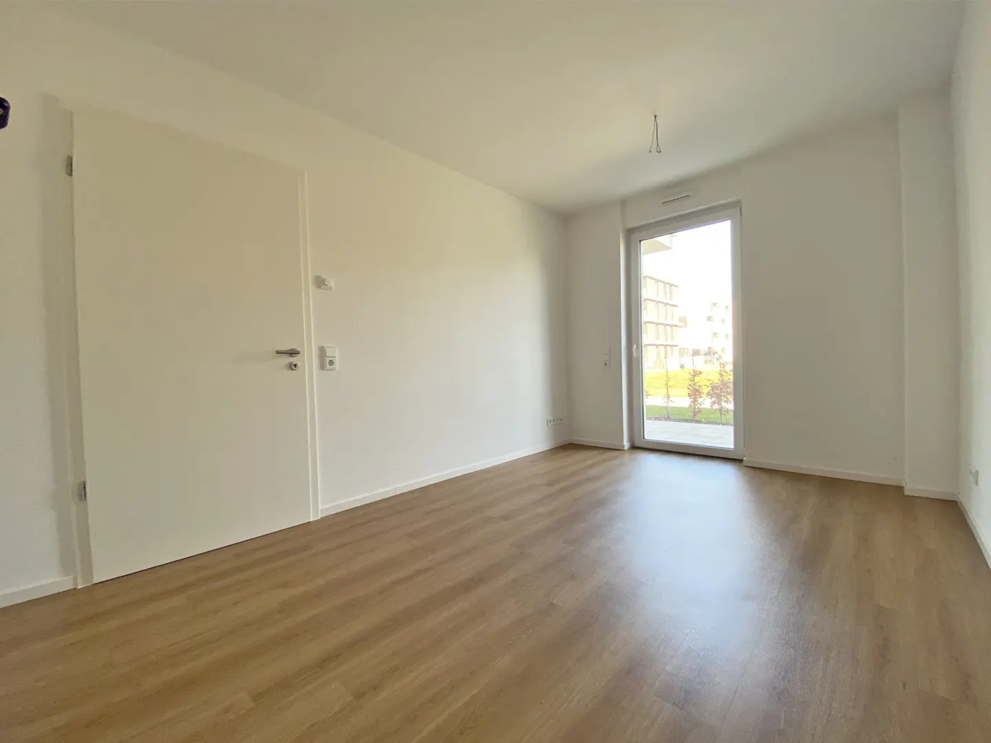 Bright, empty room with light wood floors, white walls, and a sliding glass door to a balcony. A white door is on the left.