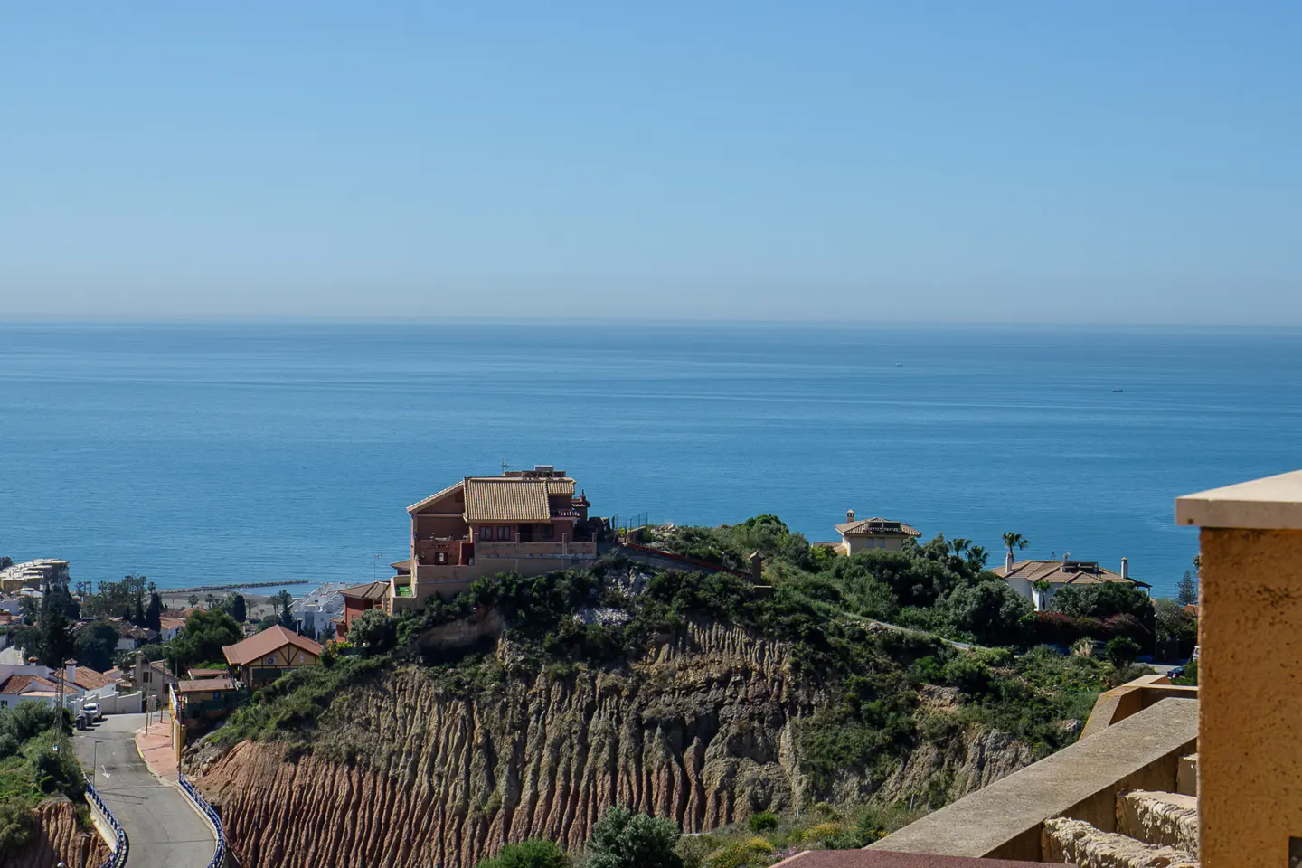 View of a brown house on a hill overlooking the blue ocean under a clear sky.