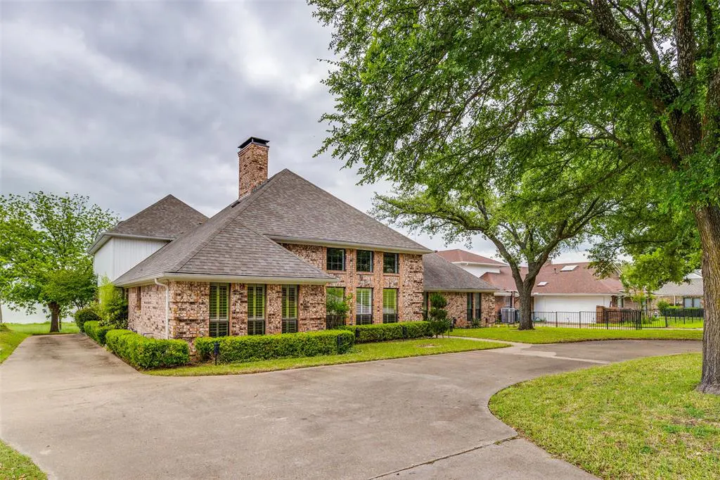 Brick house with a gray roof, chimney, and green shutters. A concrete driveway leads to the house, surrounded by green grass and trees.