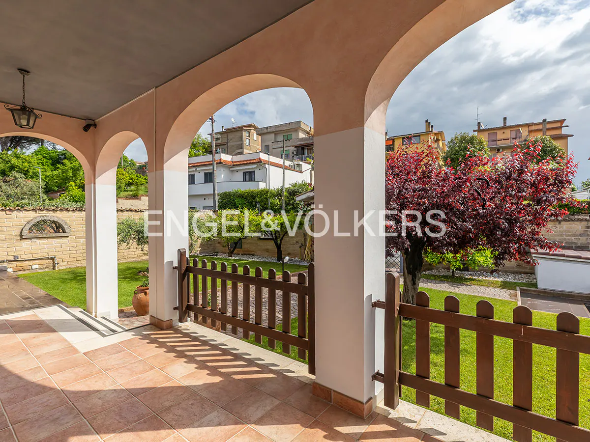 Covered patio with arched openings, tiled floor, and a brown picket fence overlooking a green lawn and trees. Engel & Völkers logo visible.