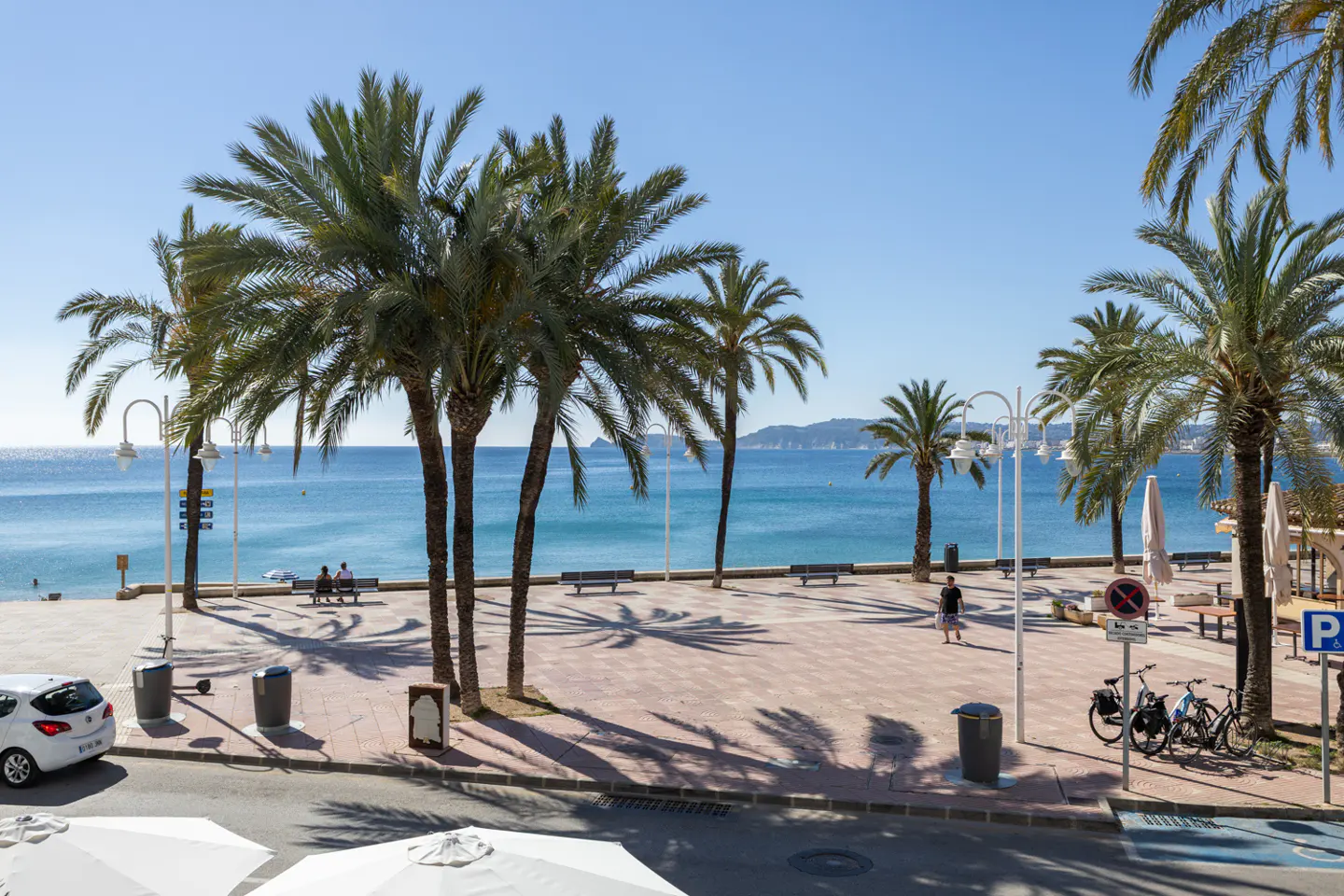 Beachfront view with palm trees, benches, and a red brick promenade under a clear blue sky. Bicycles are parked near a parking sign.