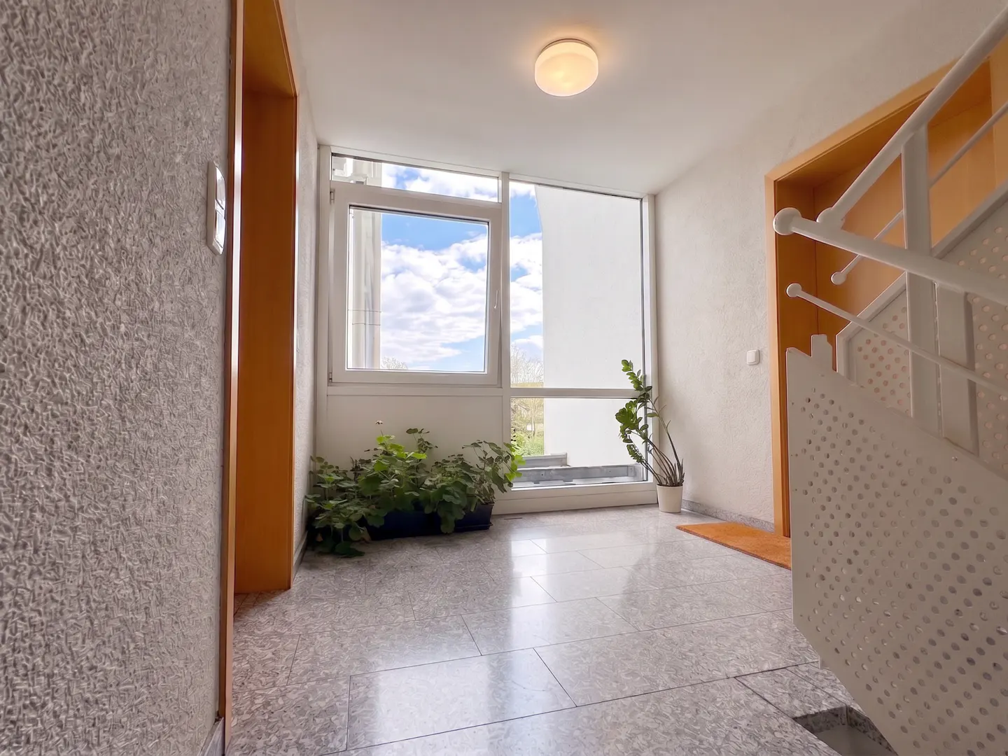 Bright entryway with light gray tiled floor, potted plants, large windows with a blue sky view, and a white staircase.