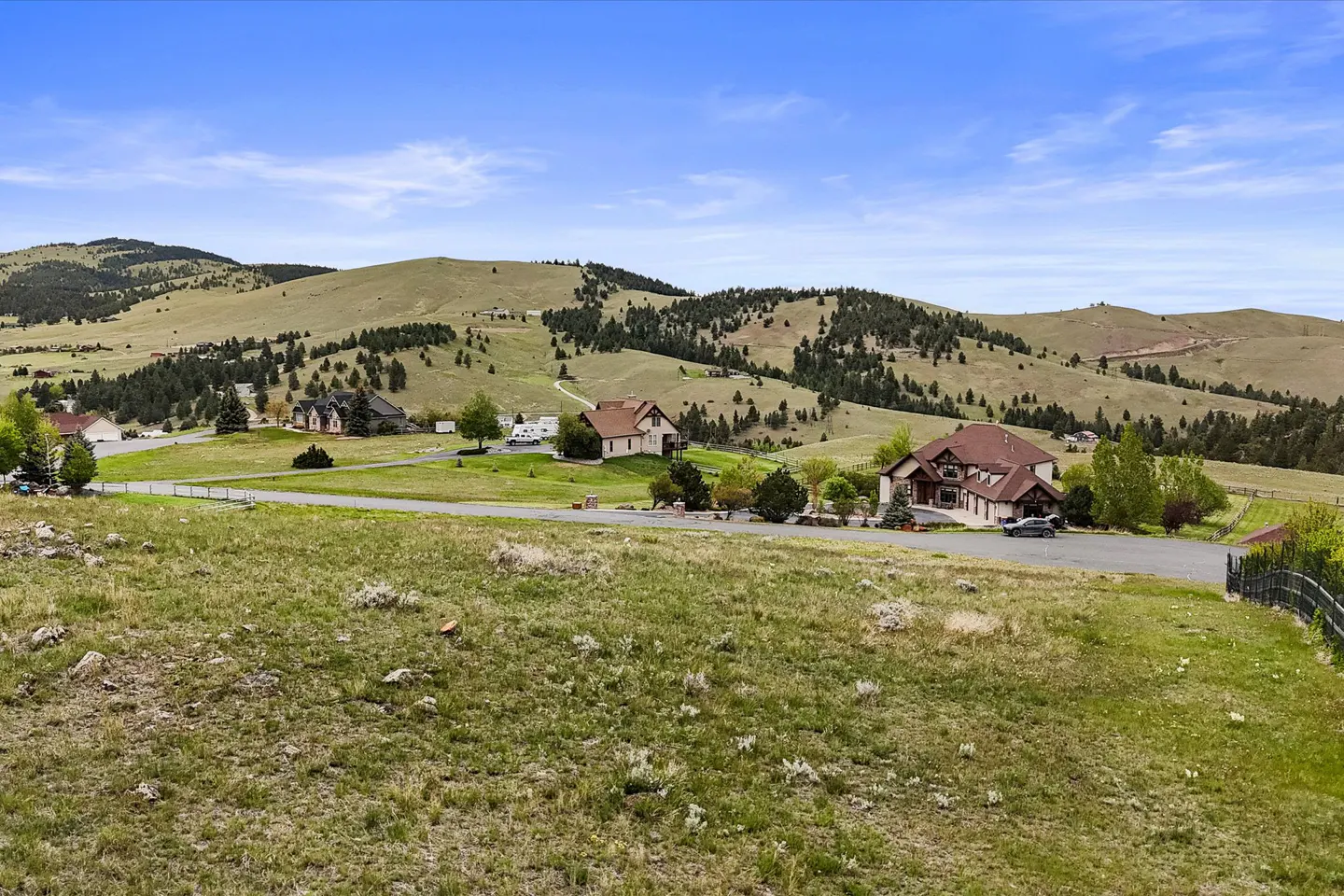 Hills with scattered trees overlook homes on grassy lots under a blue sky with clouds.