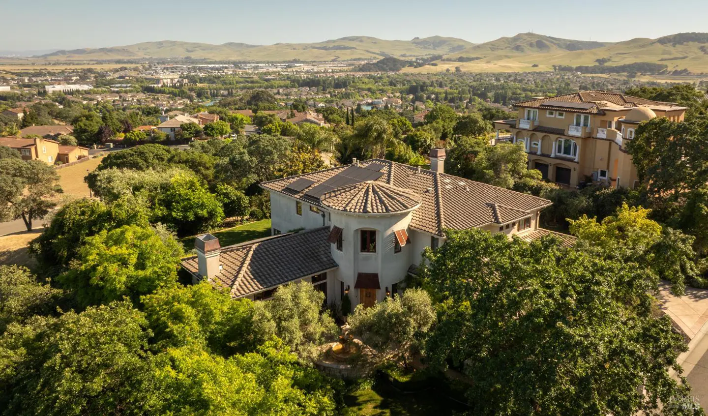 Aerial view of a large, light-colored house with a turret roof, surrounded by green trees, with mountains in the background.