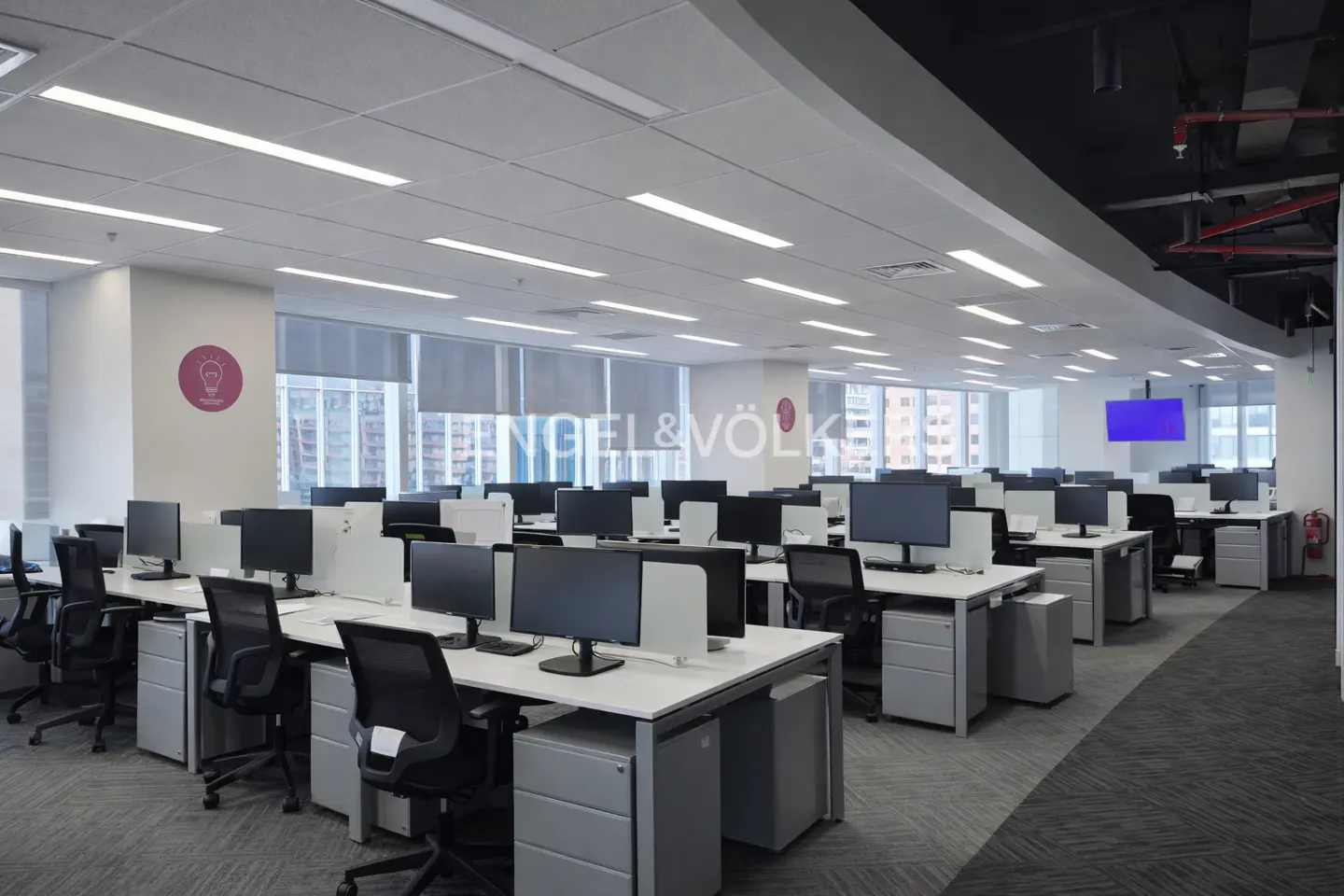 Bright, modern office space with rows of white desks, black chairs, and computer monitors. Large windows and gray carpet. Engel & Völkers logo visible.