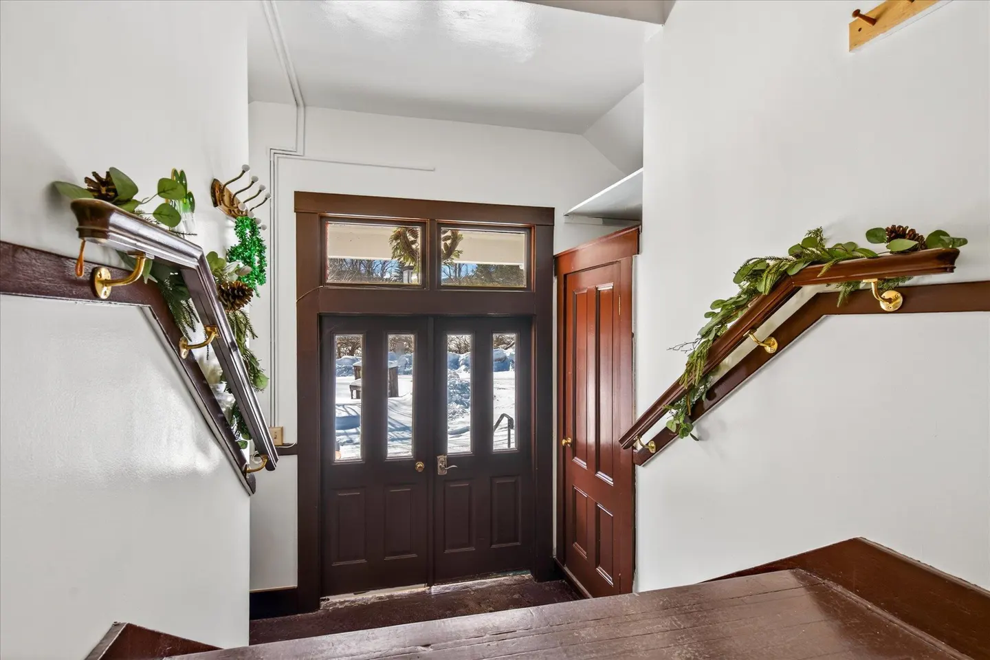 Interior view of a home's entryway with dark wood door and staircase. Garland decorates the staircase. Snow is visible outside.