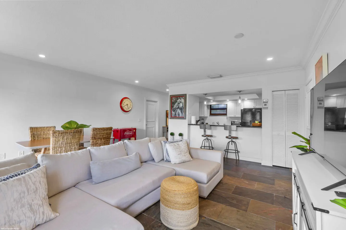 Bright, open-concept living space with a large, light gray sectional sofa, a dining table with wicker chairs, and a kitchen with a breakfast bar.