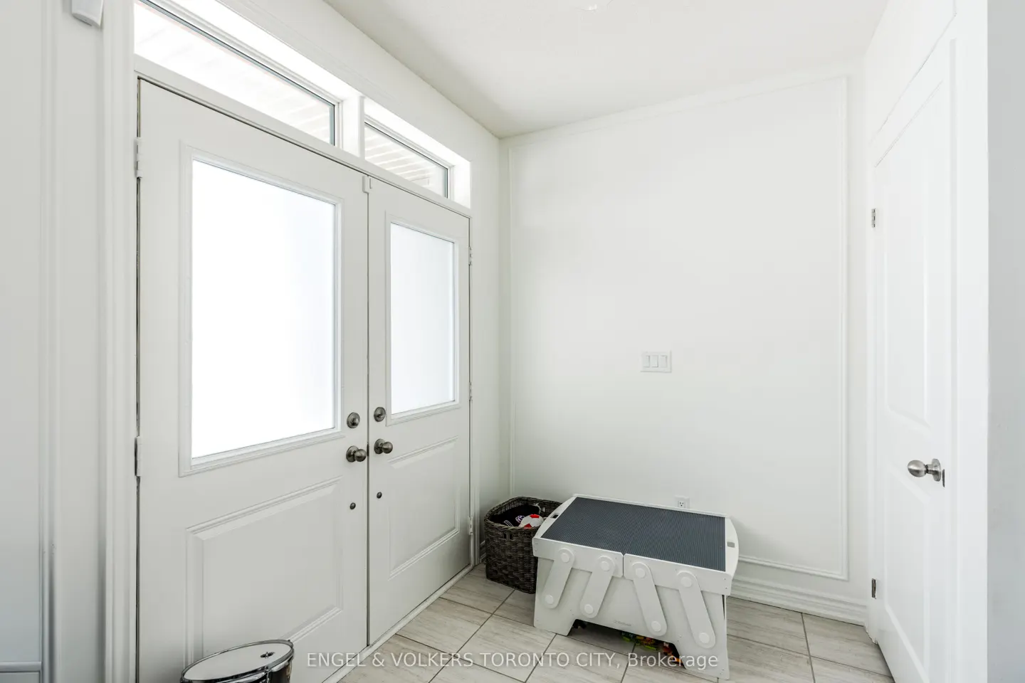 Bright entryway with white double doors, transom windows, and a white interior door. A gray step stool and basket sit on the tiled floor.