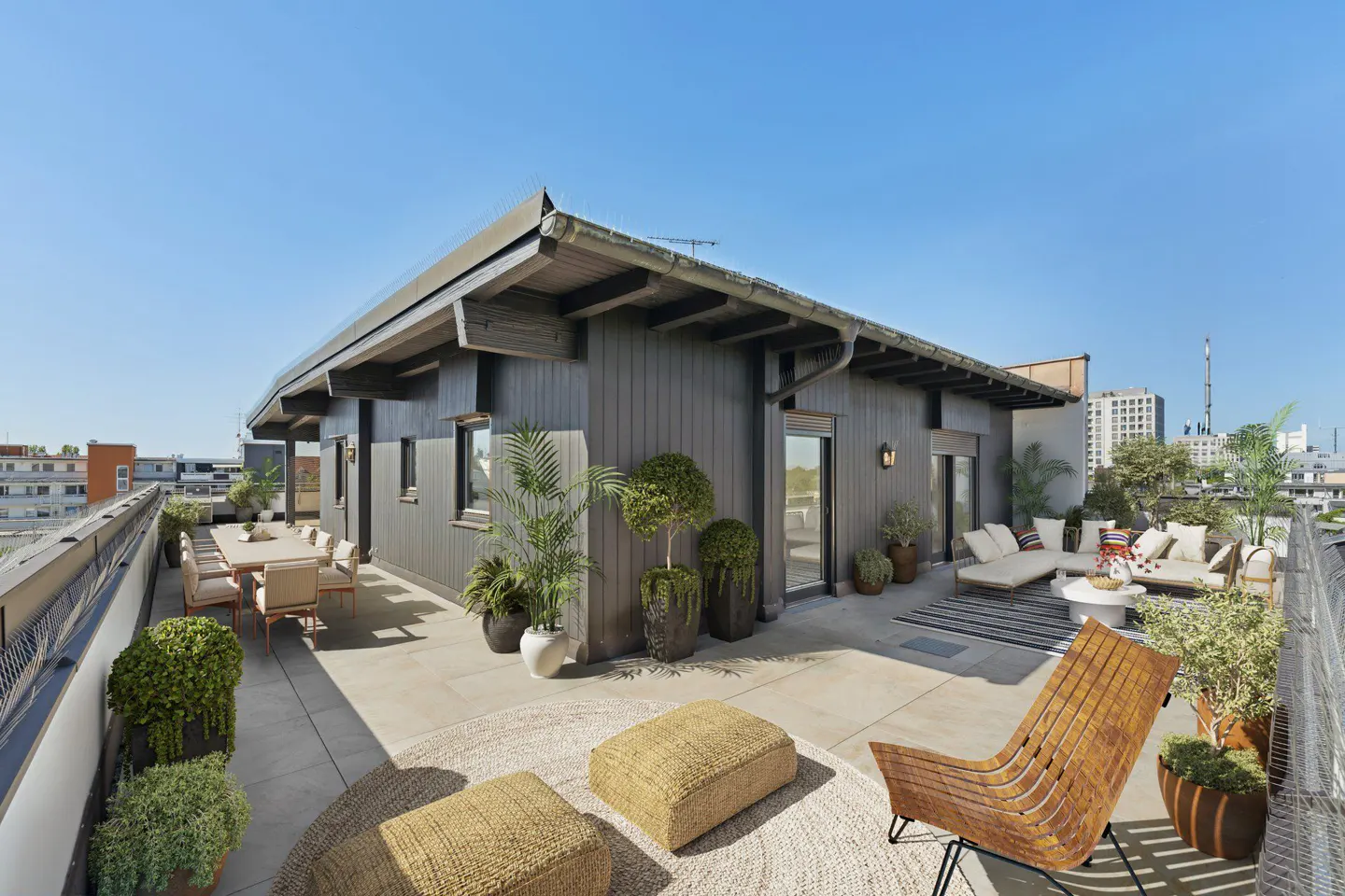Spacious rooftop terrace with dark wood siding, outdoor dining area, lounge seating, and potted plants under a clear sky.