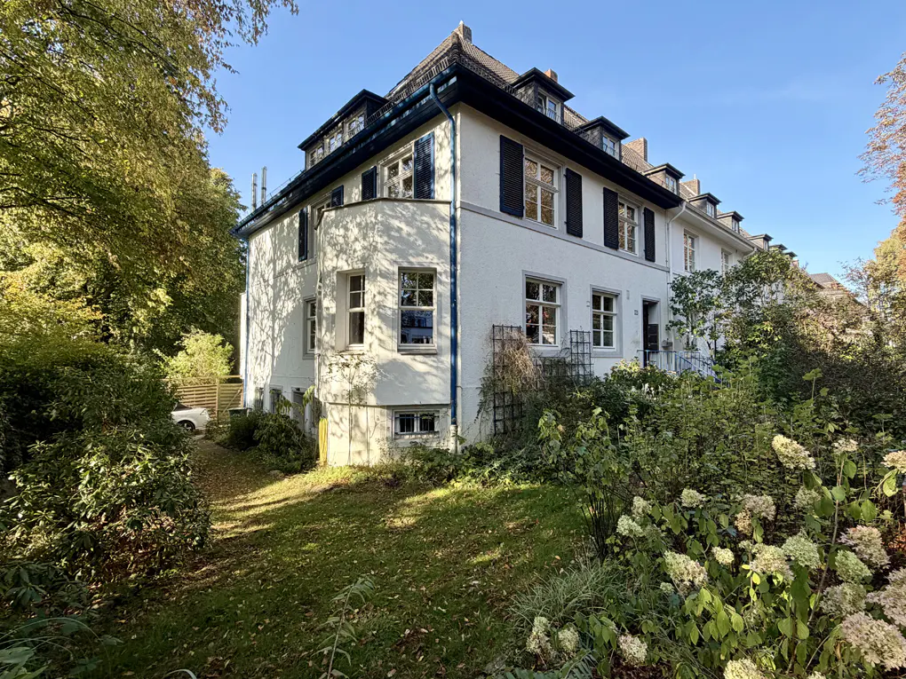 Exterior view of a white, multi-story house with black shutters, surrounded by green trees and lawn.
