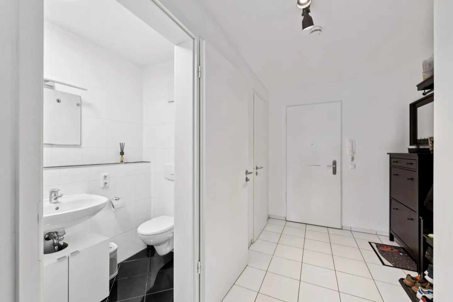 Bright hallway with white walls and tiled floor. A bathroom is visible through an open door. A dark shoe cabinet stands near the entrance.