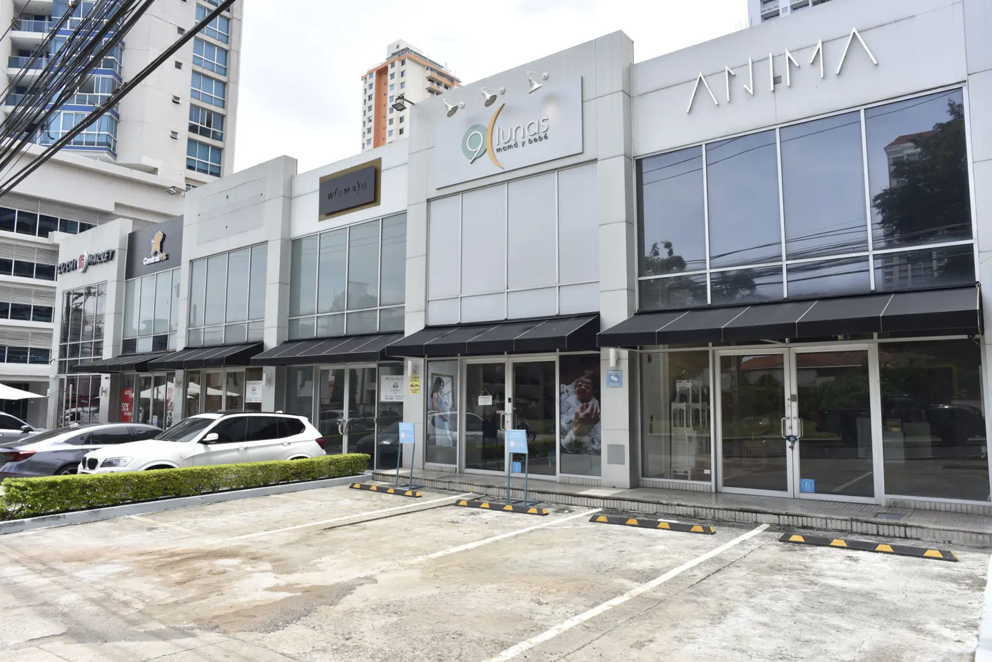 Exterior view of a modern, white commercial building with storefronts and parking. Cars are parked in front.