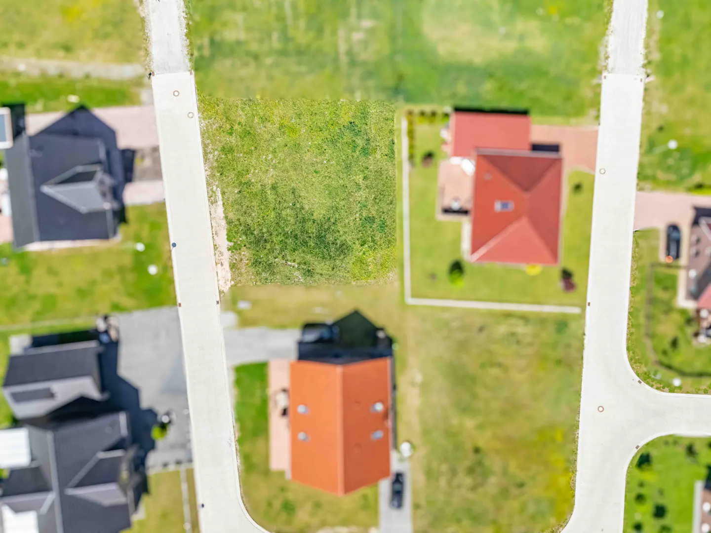 Aerial view of a vacant rectangular grass lot situated between two suburban houses with red roofs and paved roads.