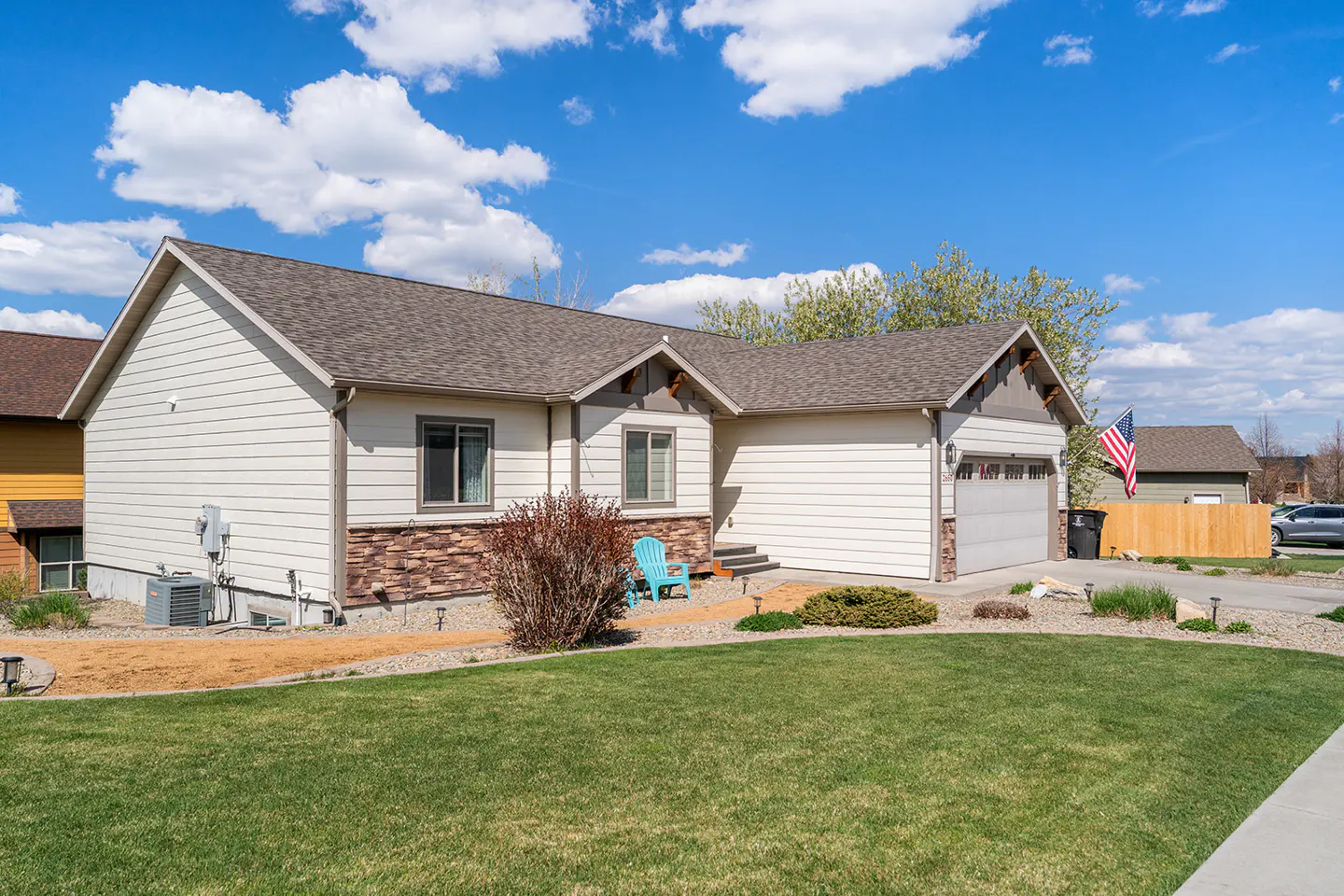 Beige single-story house with a brown roof, stone accents, and a green lawn under a blue sky with white clouds.
