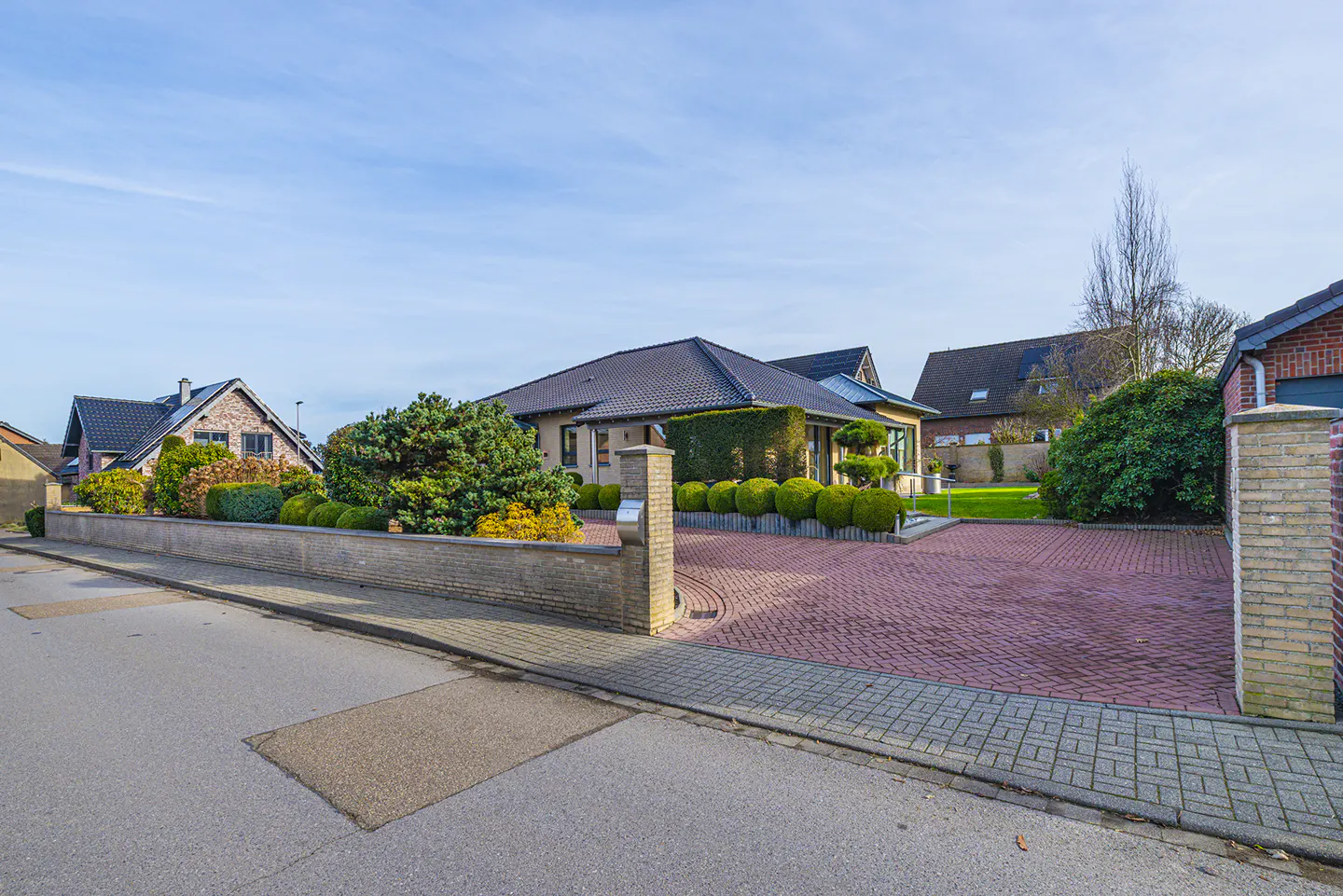A single-story house with a brick driveway and manicured green bushes under a blue sky.