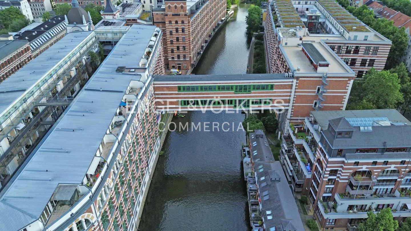 Aerial view of red brick industrial buildings connected by a green glass bridge over a narrow canal in a city.