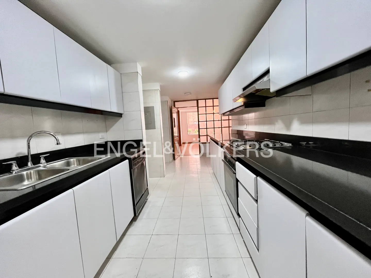 A long, narrow kitchen with white cabinets, black countertops, and white tiled floors. Appliances are stainless steel. A doorway is visible at the end.