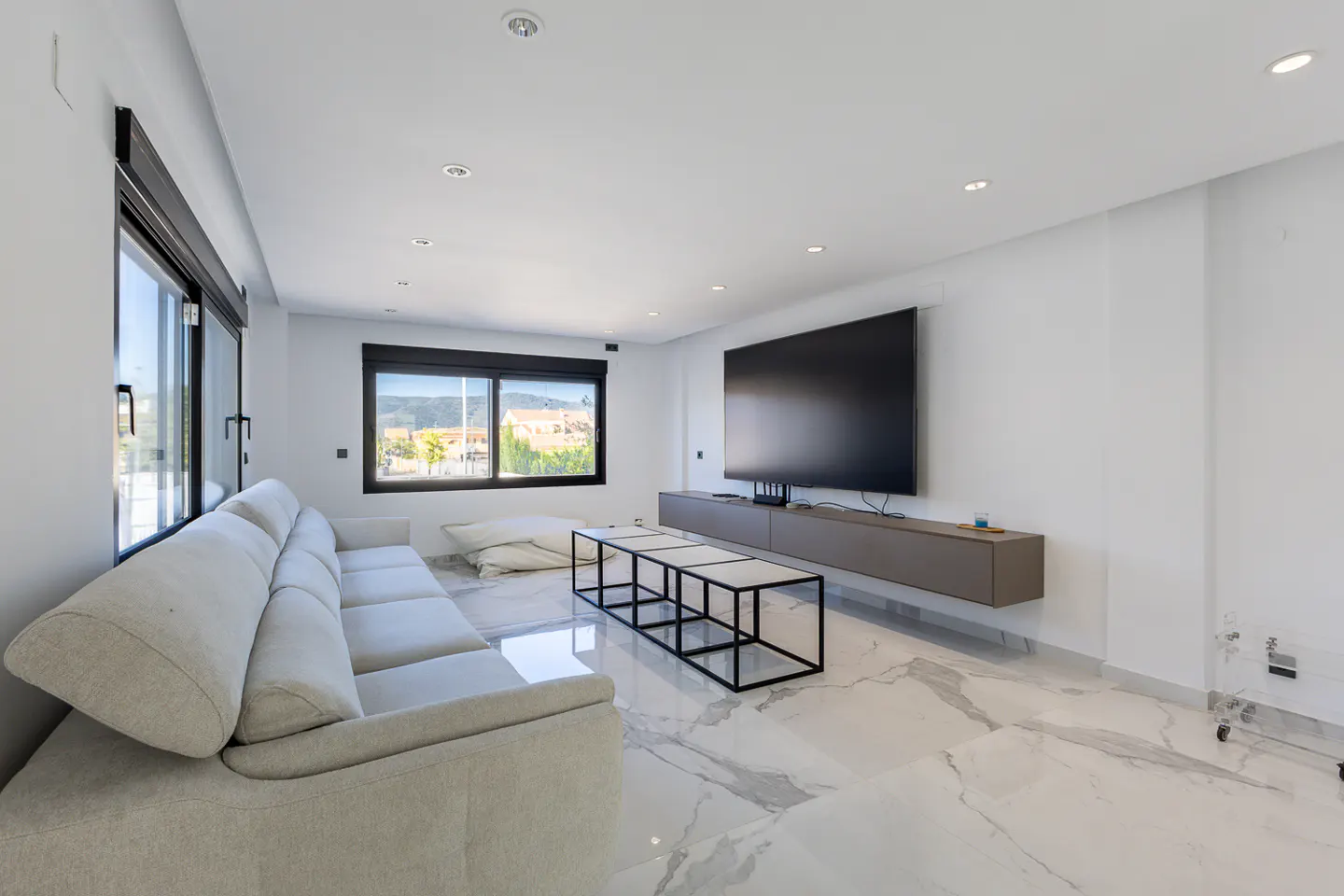 Bright living room with a beige sofa, black-framed windows, and a large TV. A black-framed coffee table sits on a marble floor.