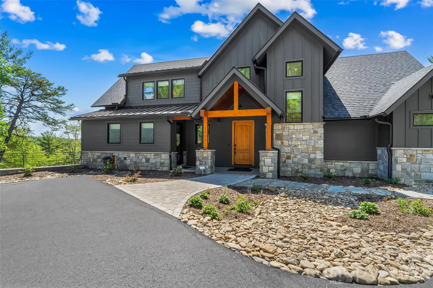 Modern gray house with stone accents, wood door, and timber porch supports. Driveway and landscaping in front. Blue sky.