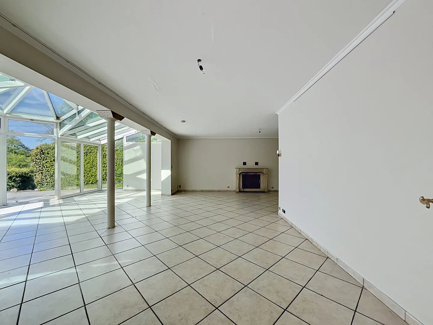 Bright, empty room with white walls, tiled floor, and a glass-enclosed sunroom area with green foliage visible outside. A fireplace is on the back wall.