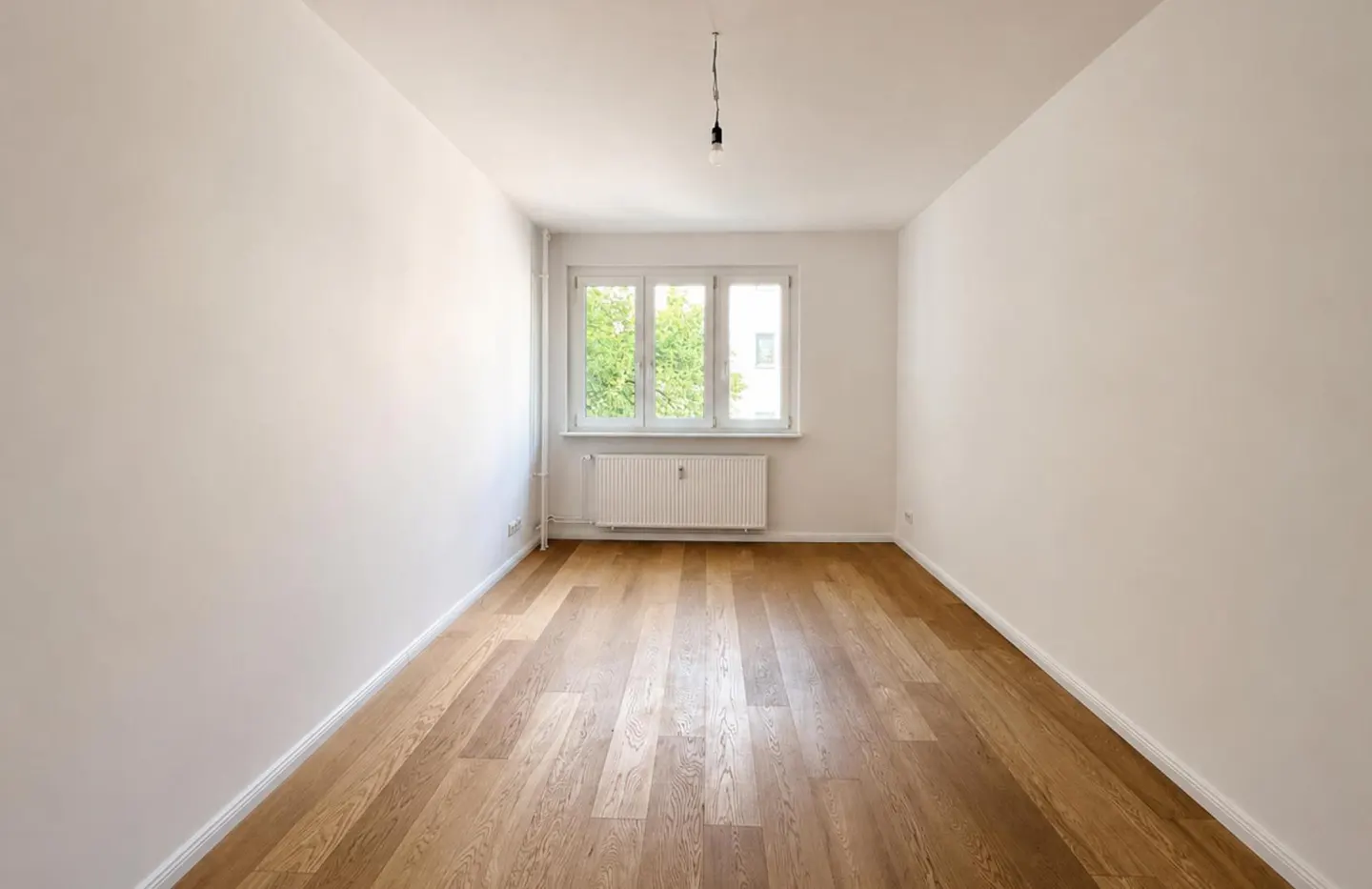 Empty room with white walls, light wood flooring, a central window, and a white radiator beneath it.