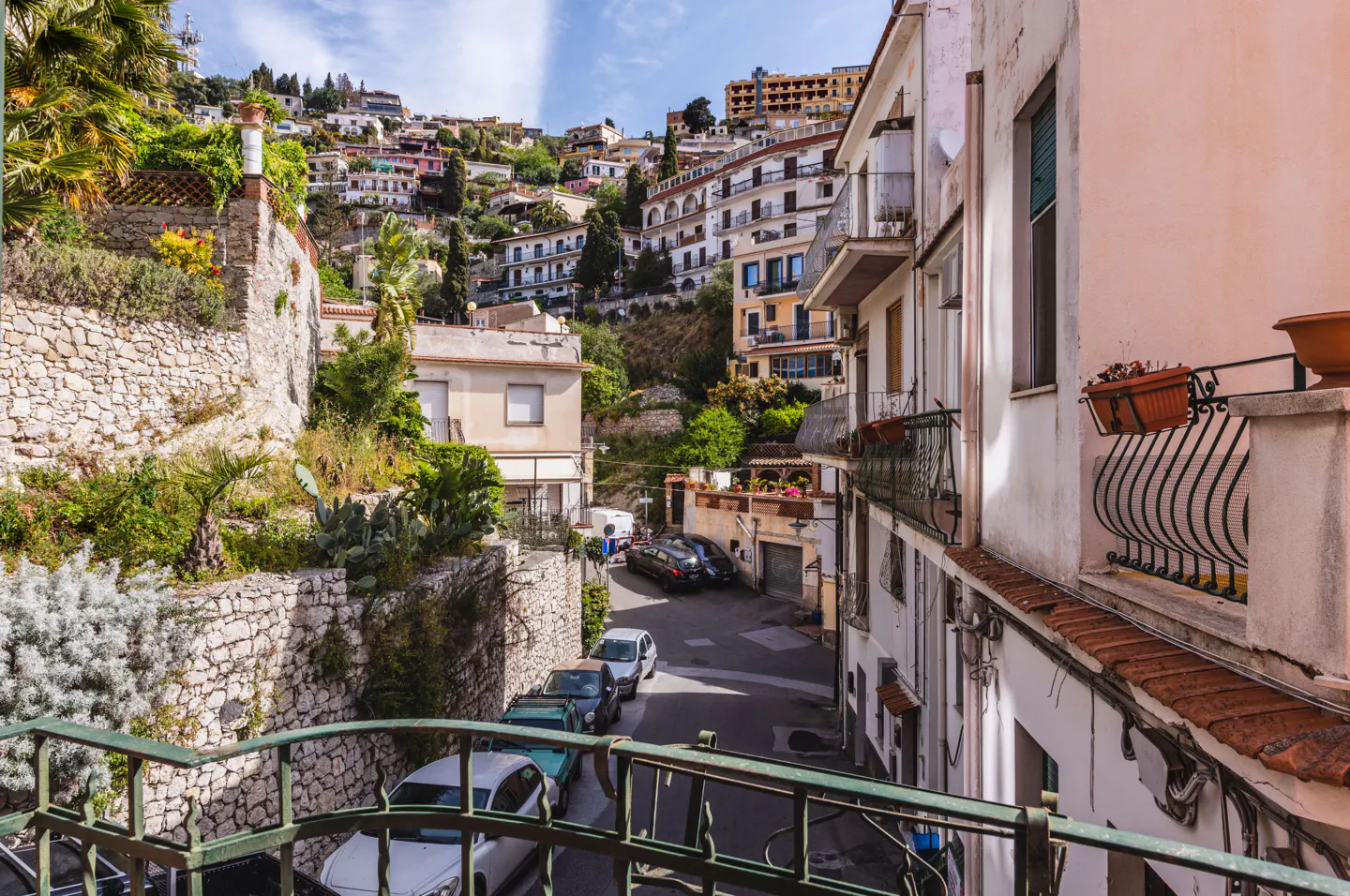 View from a balcony overlooking a narrow, sunlit street in a Mediterranean town with hillside buildings and parked cars.