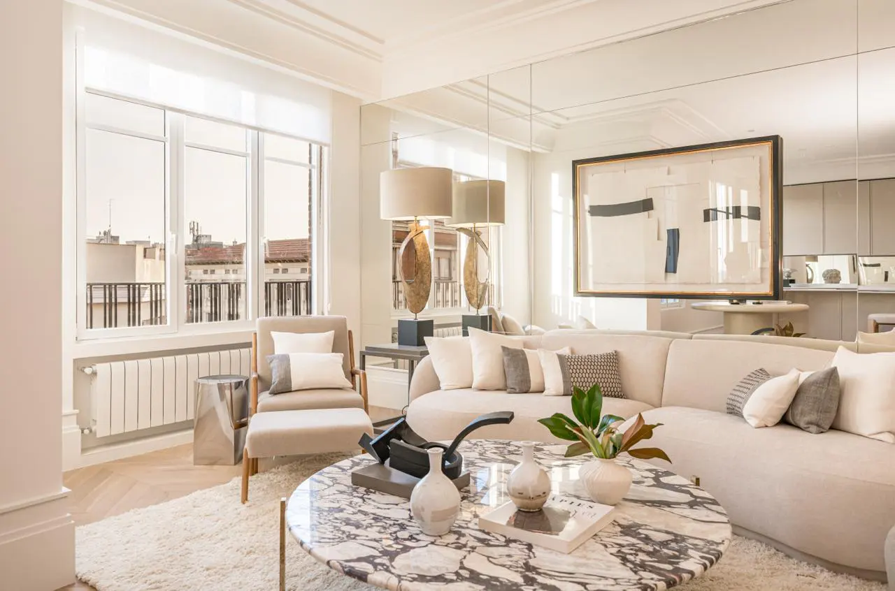 Bright living room with a beige sectional sofa, marble coffee table, and large windows overlooking city buildings. A modern art piece hangs on the wall.