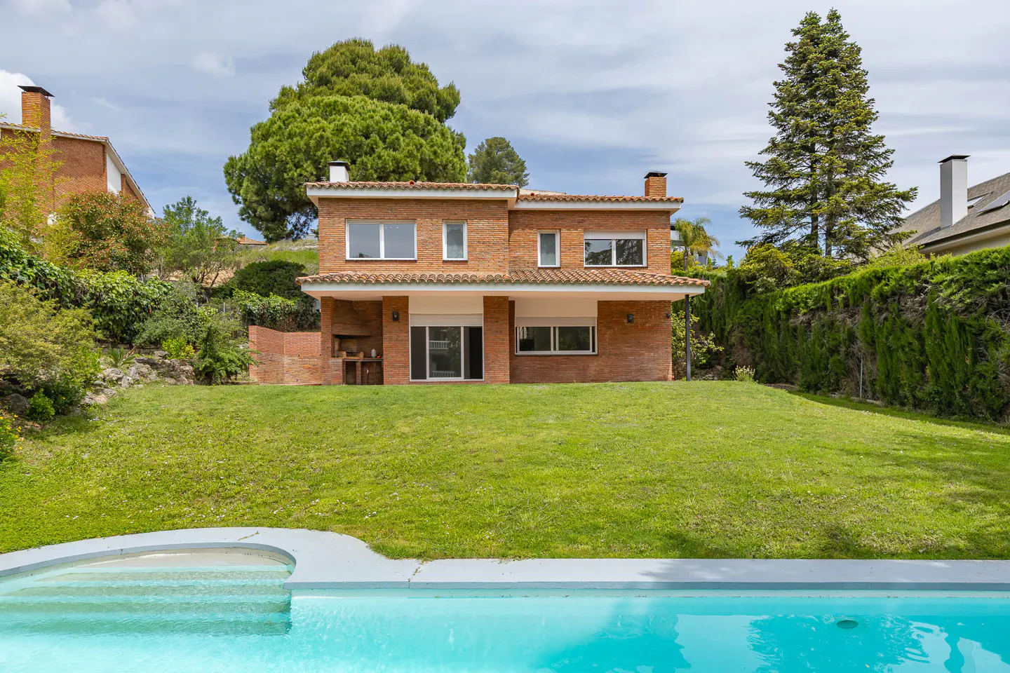 Two-story brick house with a green lawn and a blue swimming pool in the foreground.